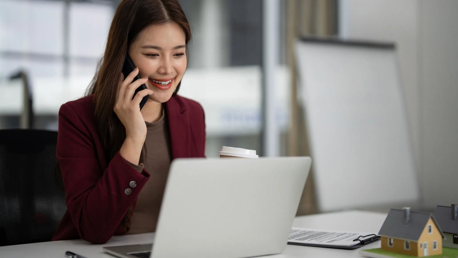Corretora de imóveis sorrindo ao falar ao telefone enquanto trabalha no notebook, com uma maquete de casa sobre a mesa